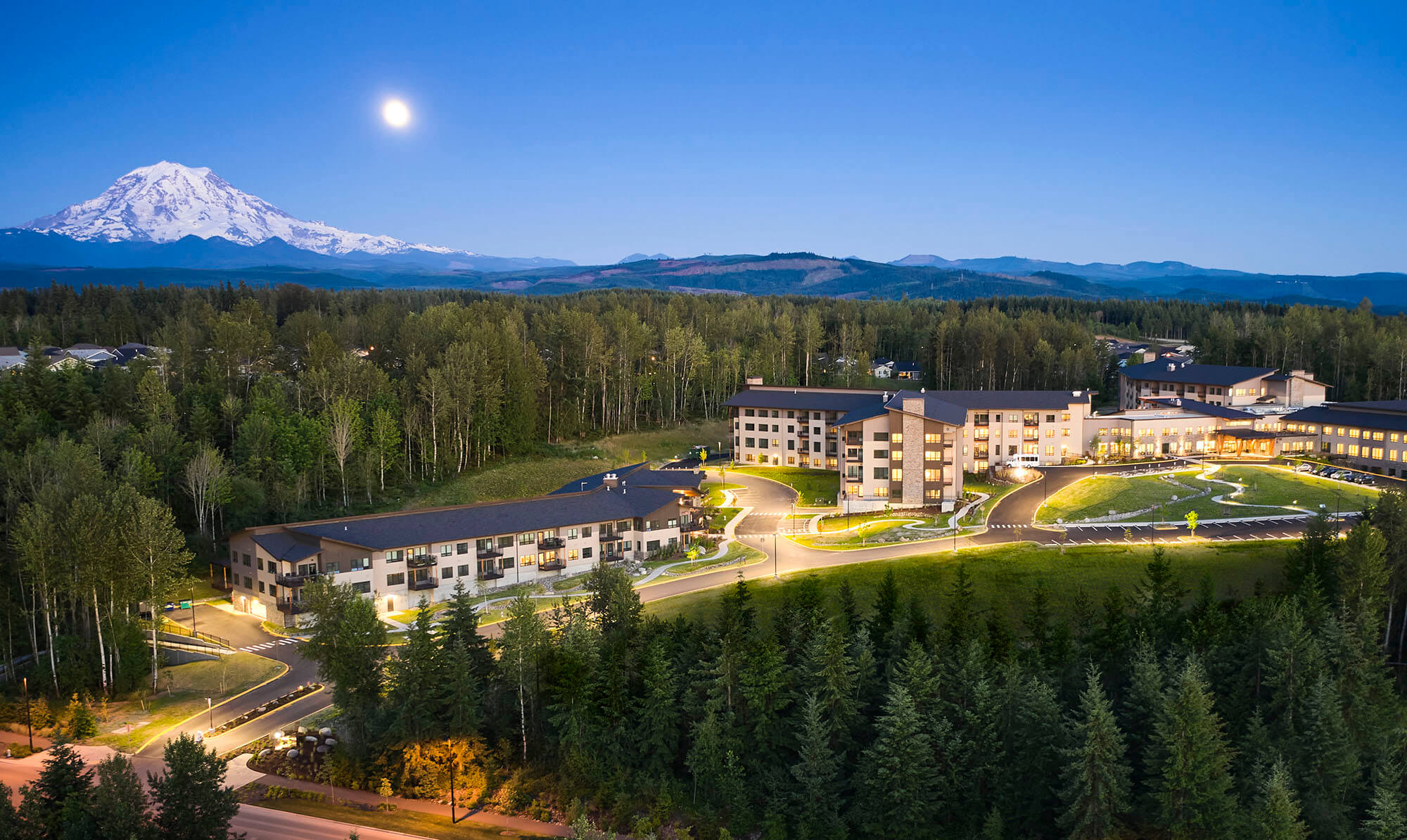 Group of buildings surrounded by pine trees with moon and Mount Rainier in the background
