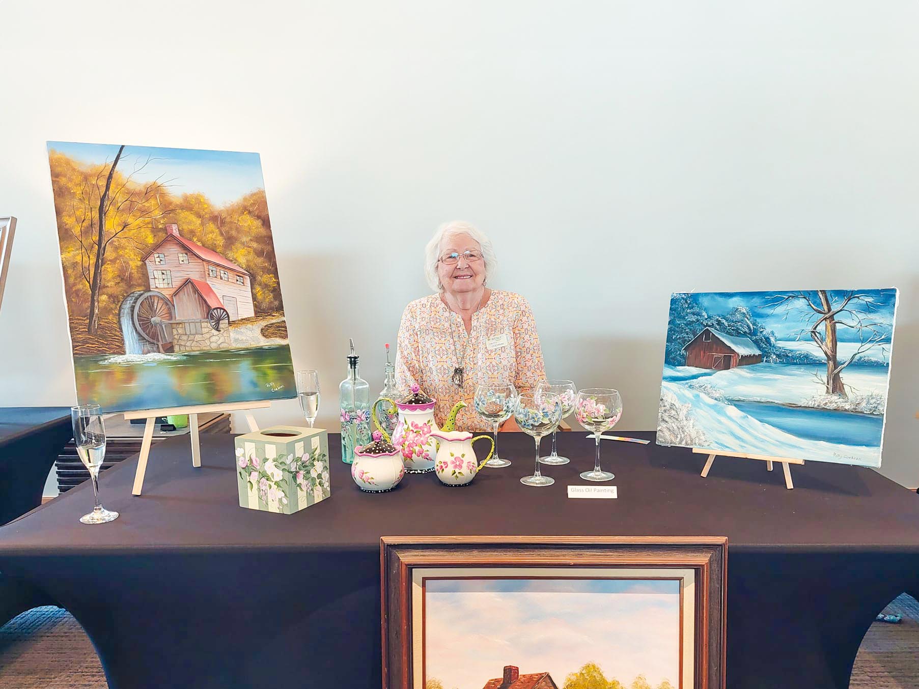 Kind and happy looking older woman with paintings and sculpture art around her seated at a table