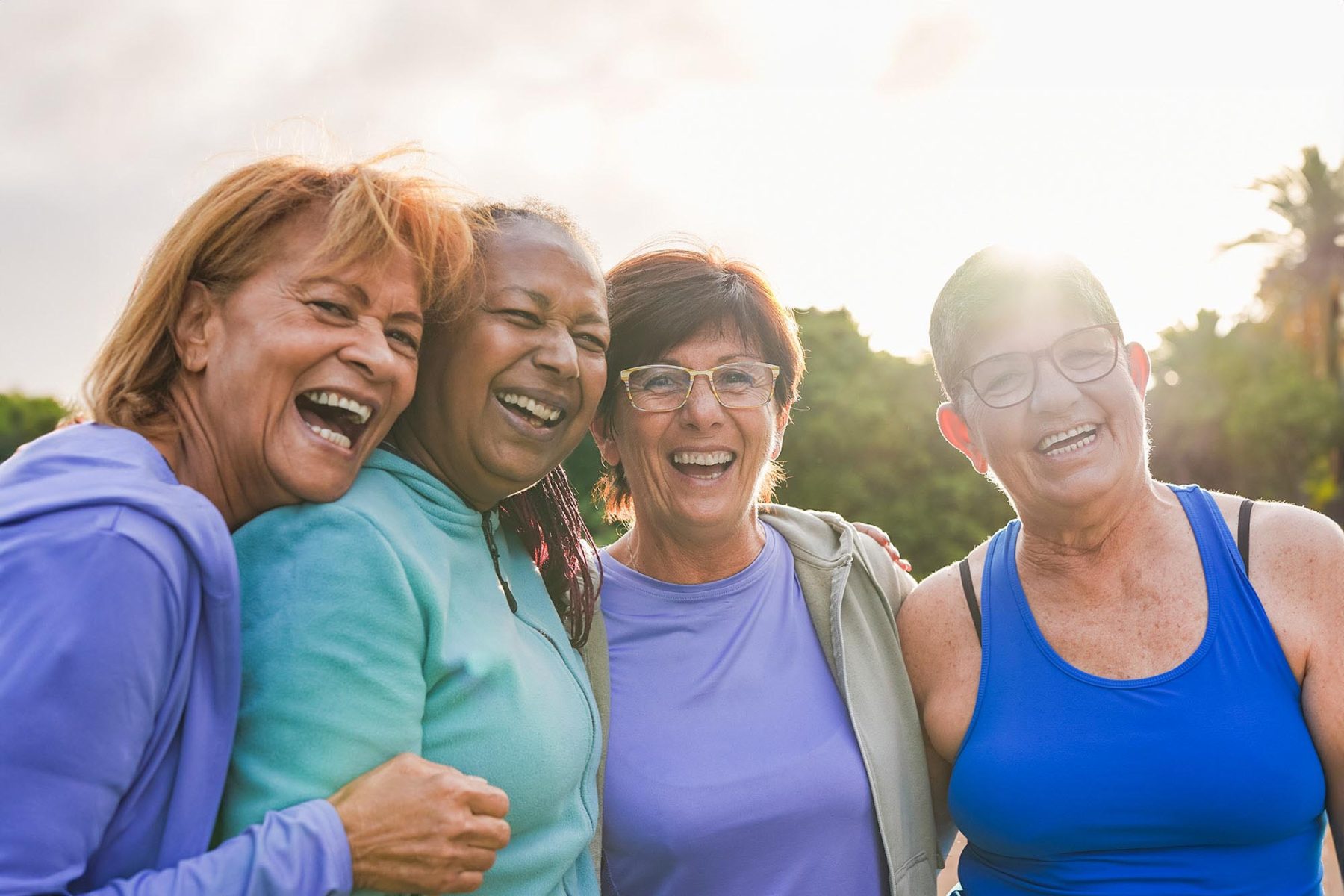 Group of senior multiracial women hugging each other after sport workout outdoor - Fit elderly female friends with sunset in background - Healthy lifestyle concept