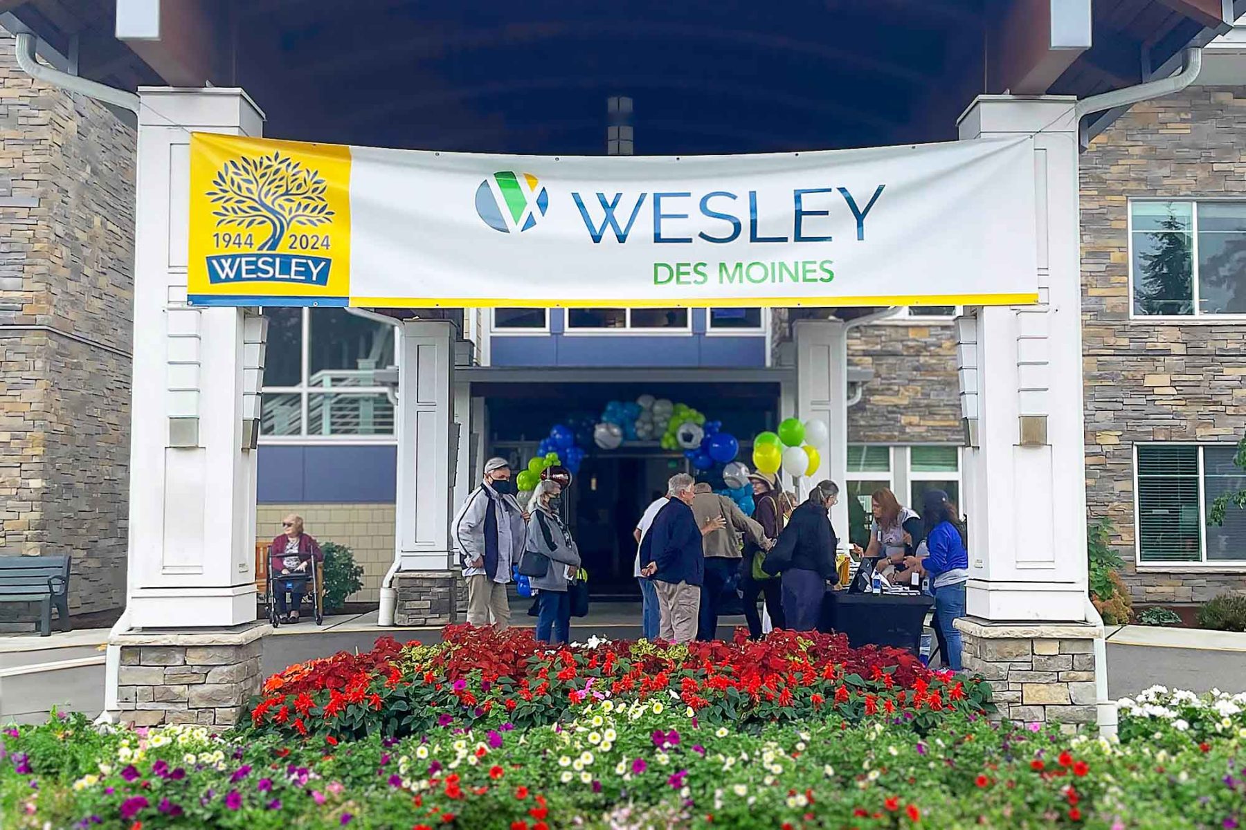 People gathering outside of the new Wesley Des Moines Gardens entrance to celebrate the dedication and completion of the campus. Colorful flowers in front with a banner commemorating the event.